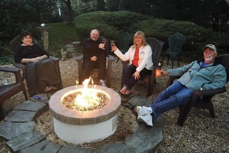 Four people around a lit fire pit in a backyard. One woman raises a glass. Everyone sits in chairs and smiles.