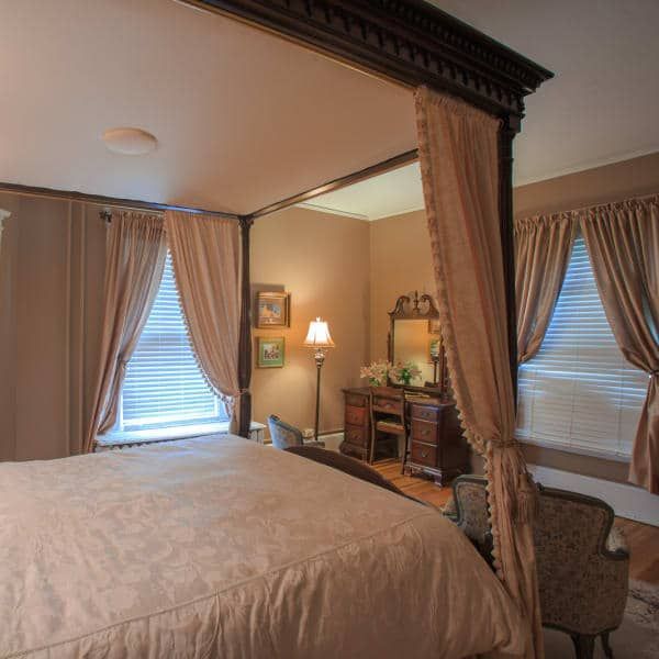 Bedroom with a large bed, curtains, windows, and a vanity. Warm tones and ornate decor create a cozy atmosphere.