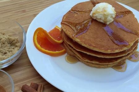 Stack of pancakes with syrup, butter, orange slices, and brown sugar on a white plate.