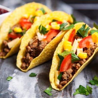 Three hard-shell tacos filled with ground meat, tomatoes, corn, cheese, and parsley, on a wooden surface.