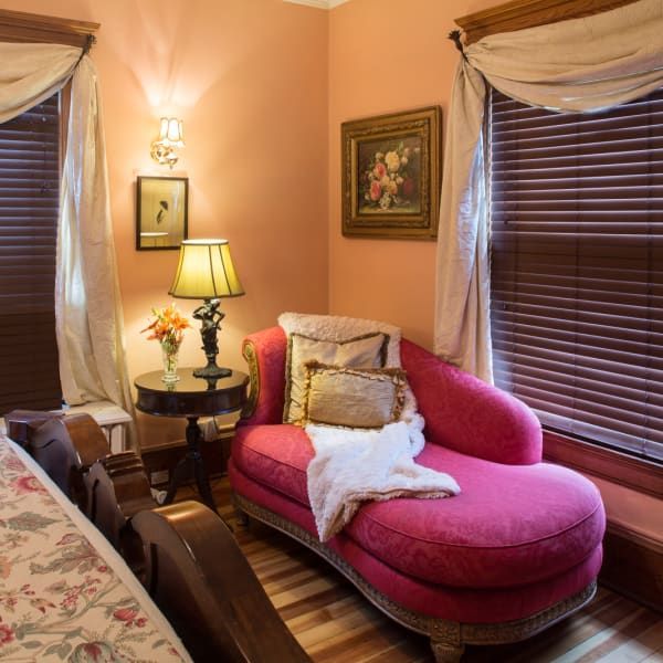 Pink chaise lounge in a peach-colored room, near a window with brown blinds and a small table with a lamp.