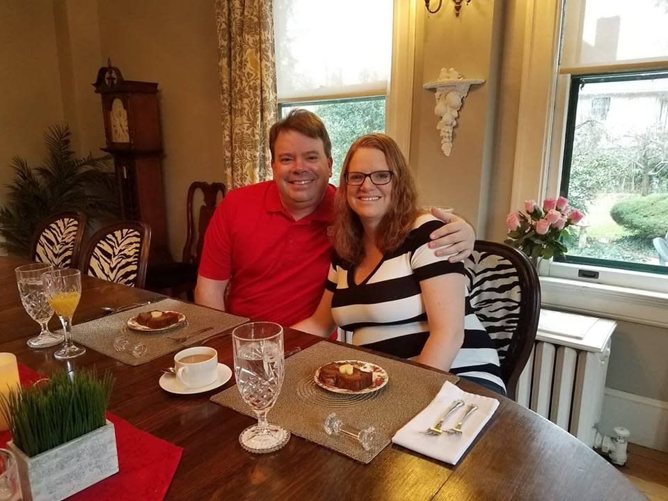 Couple at a dining table, smiling. Man in red shirt, woman in stripes. Food and drinks set before them.