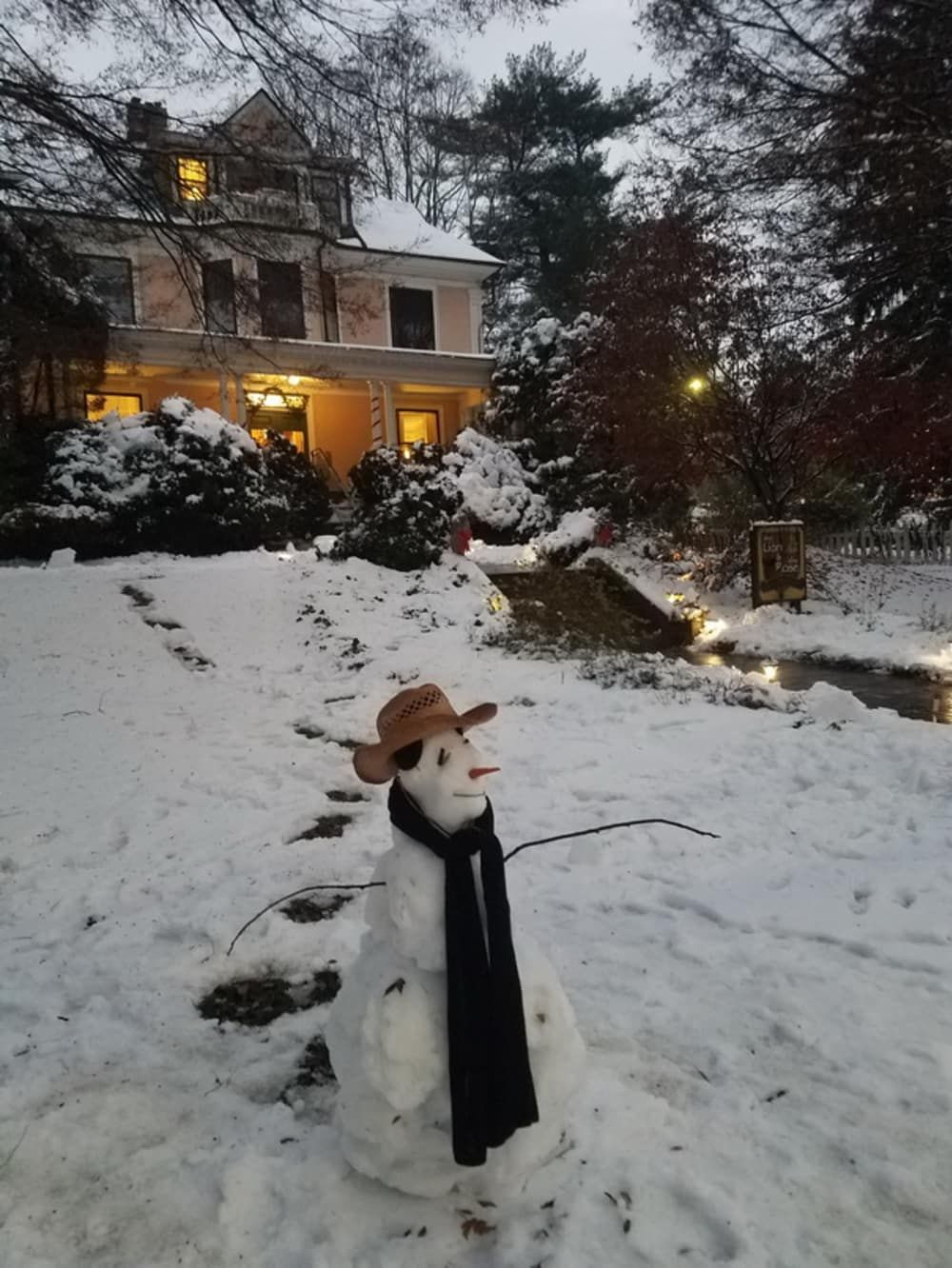 Snowman in a hat and scarf stands in front of a snow-covered house with lit windows.