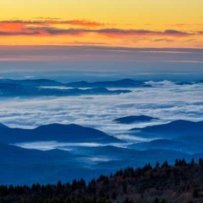 Blue mountain range at dawn, topped by fog, under orange and yellow sky.