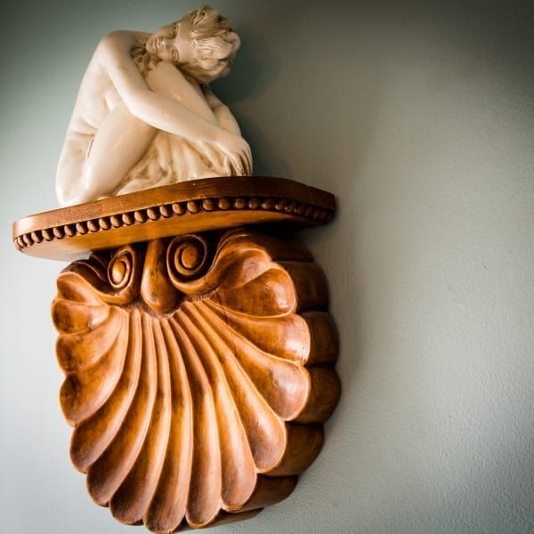 Carved wooden shell-shaped shelf holding a small white statue of a nude person, against a blue-green wall.