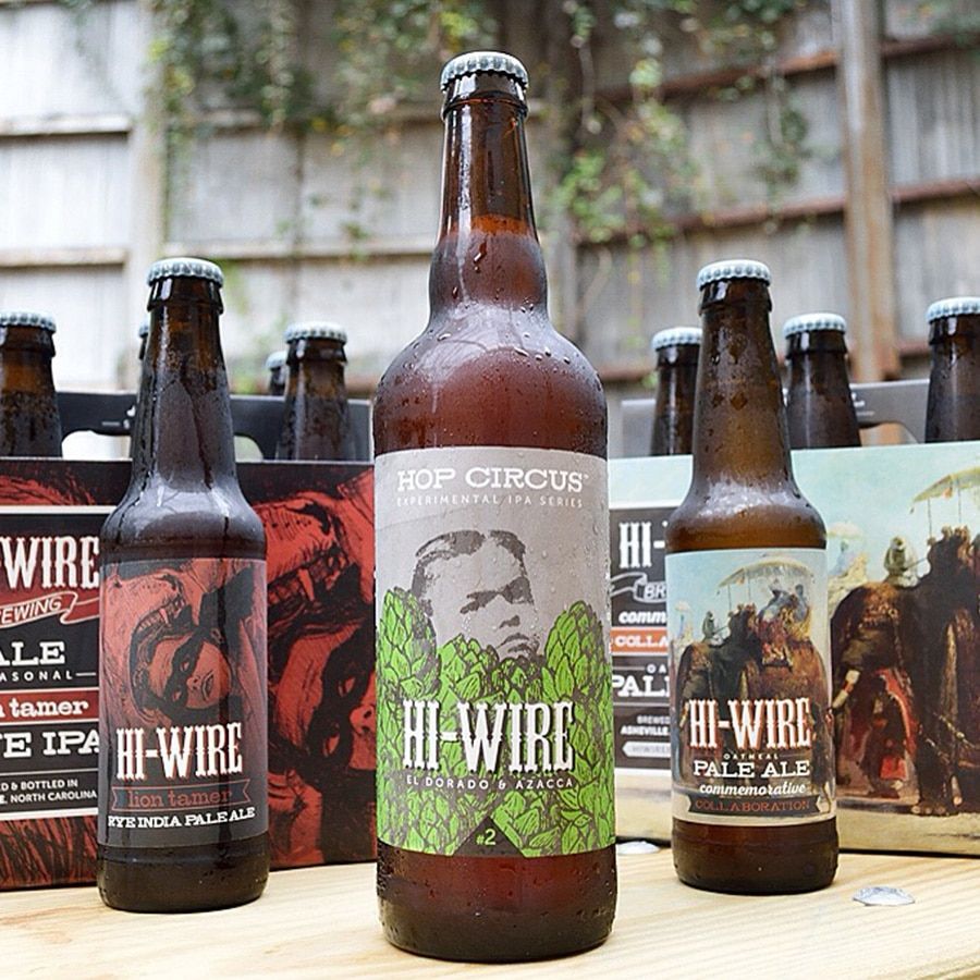 Three bottles of Hi-Wire beer with different labels, on a wooden surface, with a blurred background of more beer bottles.