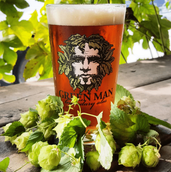 A glass of amber beer with a Green Man logo, surrounded by hops on a wooden surface.