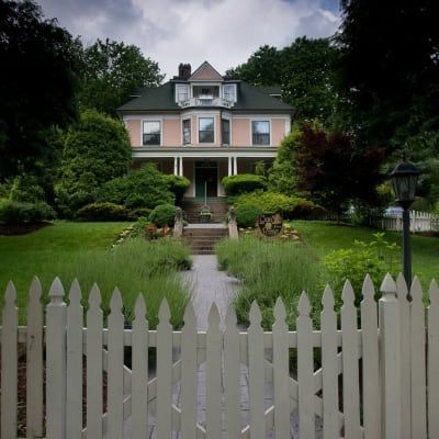 Pink house with green roof, white picket fence, pathway, and lush landscaping.