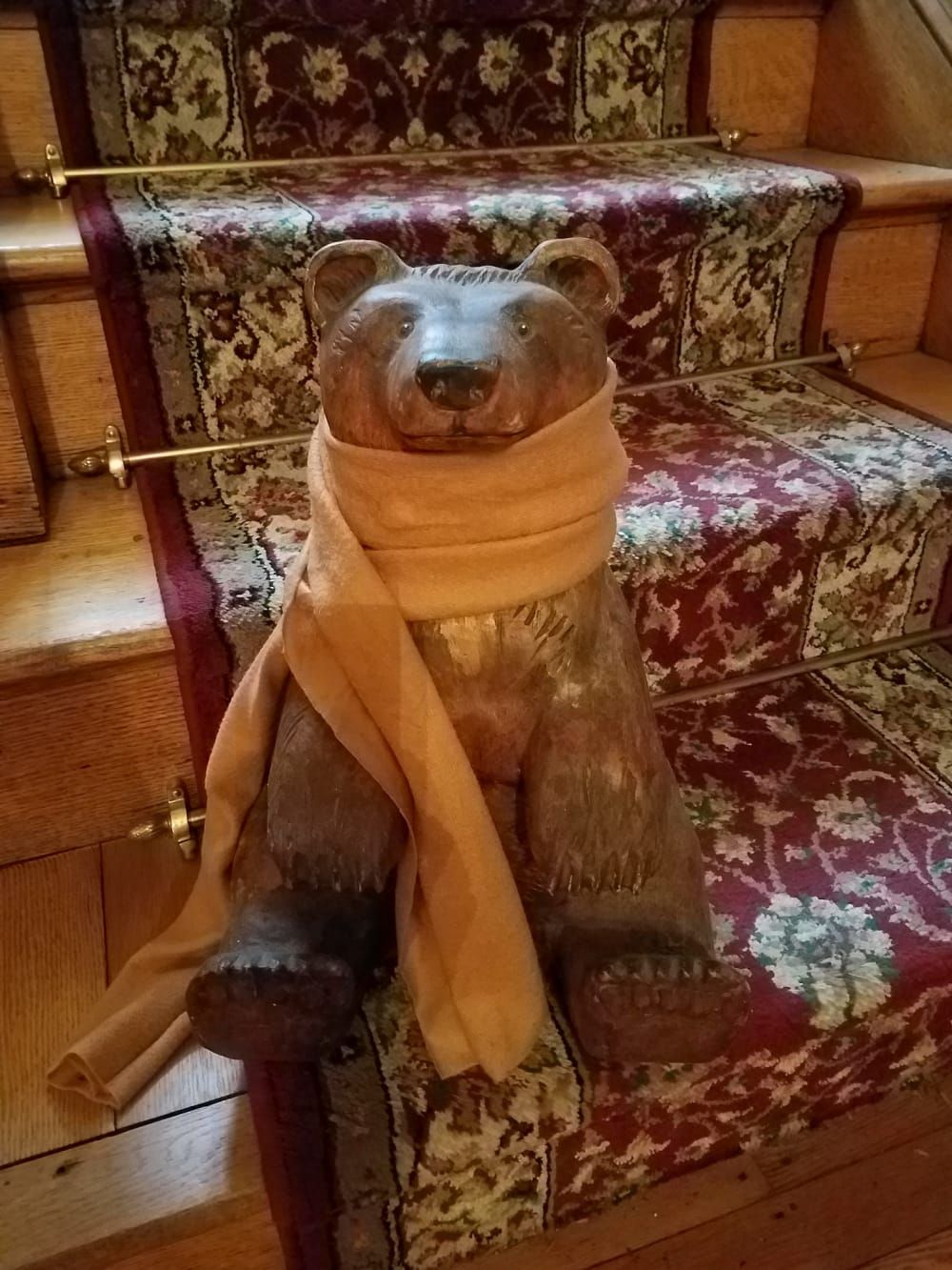 Wooden bear statue wearing a scarf, sitting on a patterned rug-covered staircase.