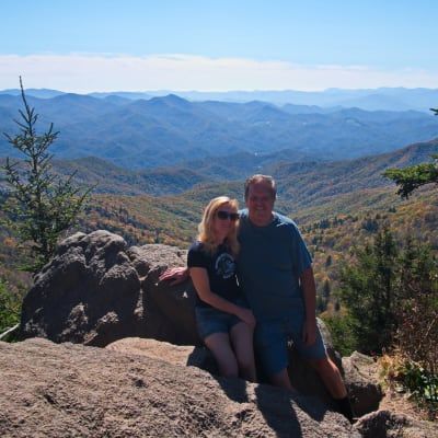 Couple poses on rocky mountaintop with mountain range in background.