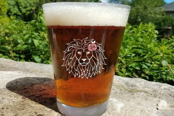 Pint glass of beer with lion logo, resting on stone wall, with greenery in background.