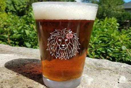 Pint glass of beer with lion logo, resting on stone wall, with greenery in background.