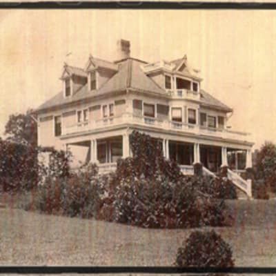 Large, light-colored Victorian-style house with multiple levels, balconies, and dormers. Front porch, surrounded by greenery.