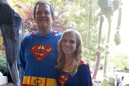 Couple in Superman costumes pose outdoors, smiling, near a skeleton decoration.