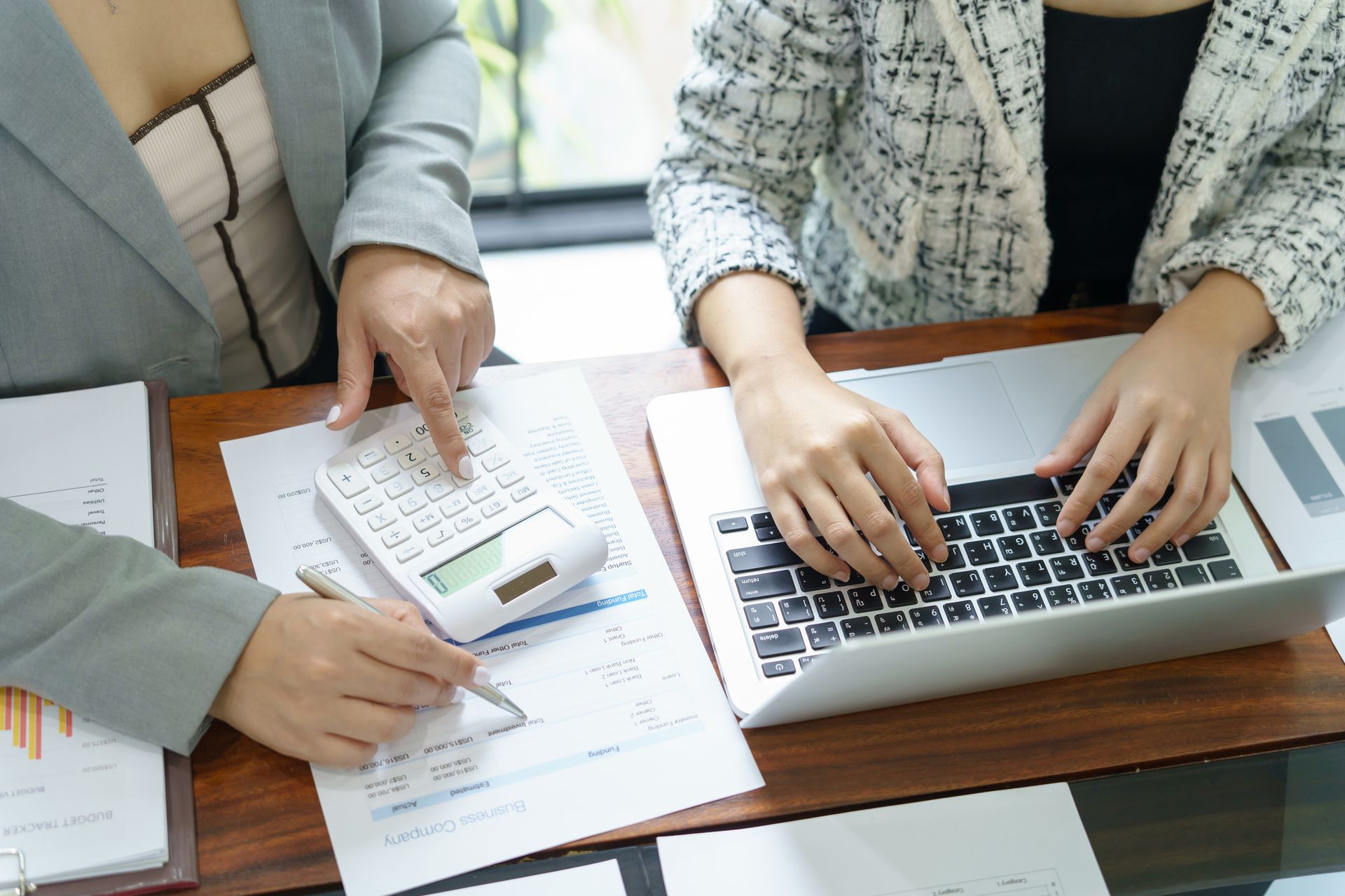 Two women are sitting at a table using a laptop and a calculator.