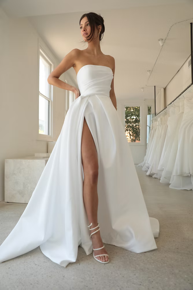 Woman in strapless white wedding dress with high slit, standing indoors, looking away.
