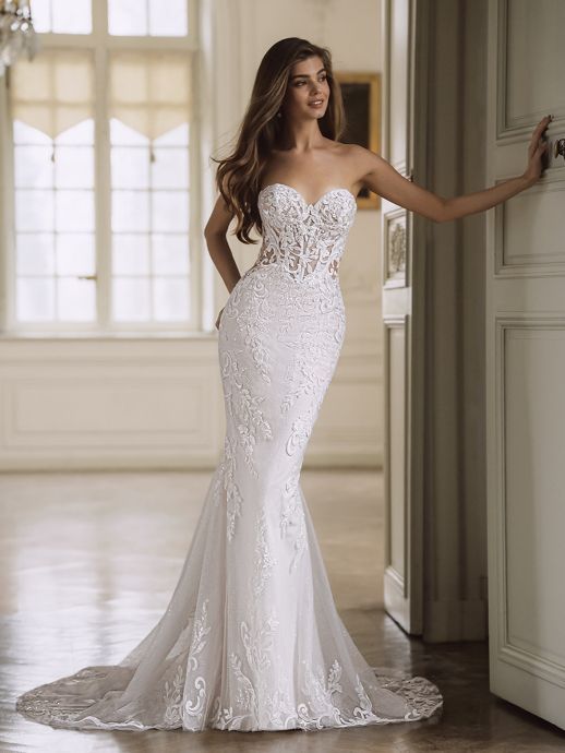 Woman in a white, strapless, lace wedding dress, posing in a bright hallway, smiling.