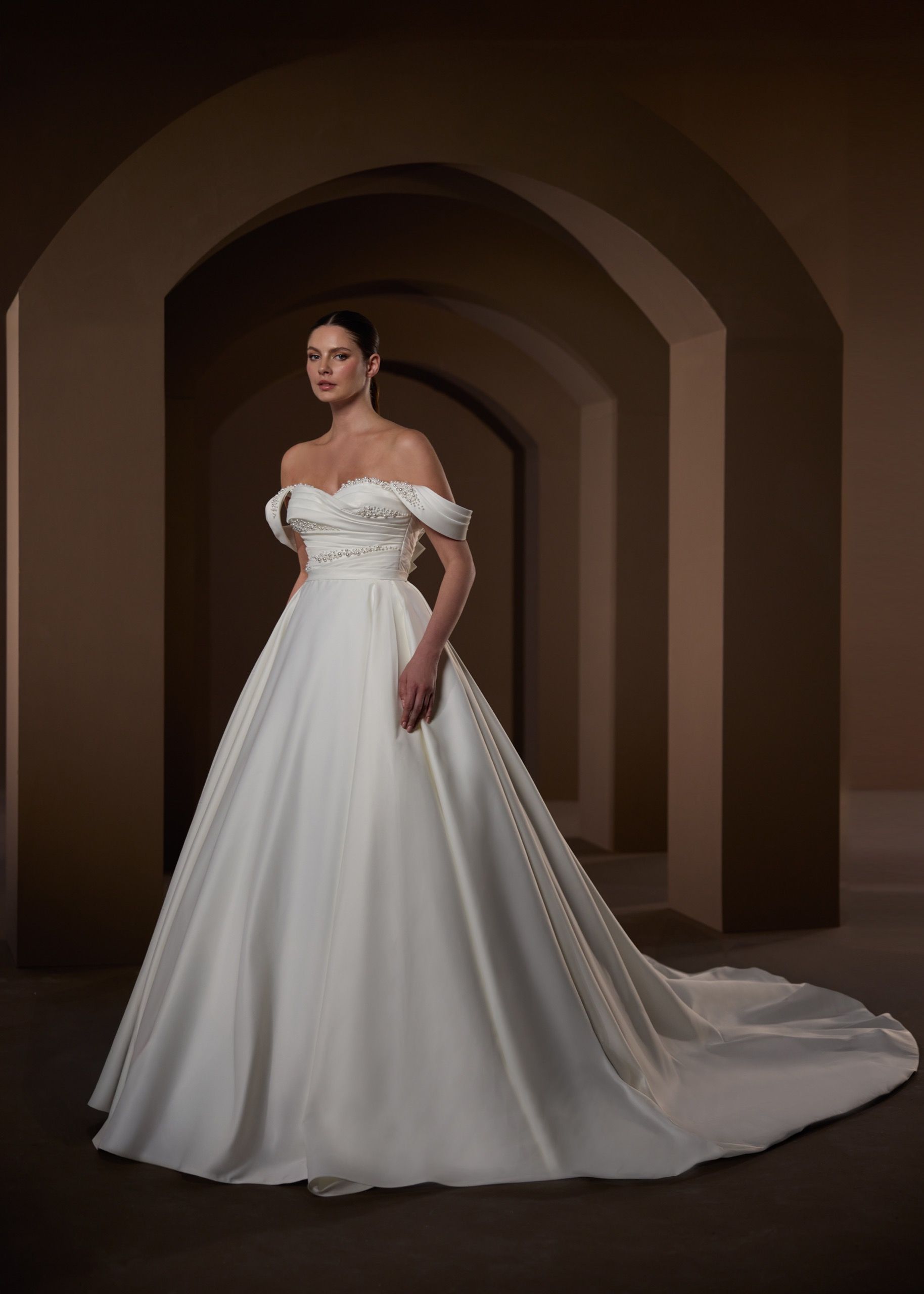 Woman in a white off-the-shoulder wedding dress with a long train, standing in an arched hallway.