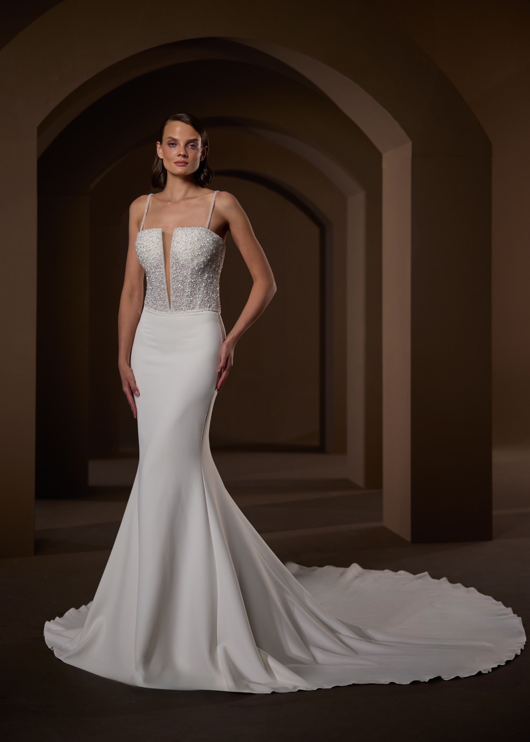 Bride in a white mermaid-style wedding gown with beaded bodice and long train, standing in an arched hallway.