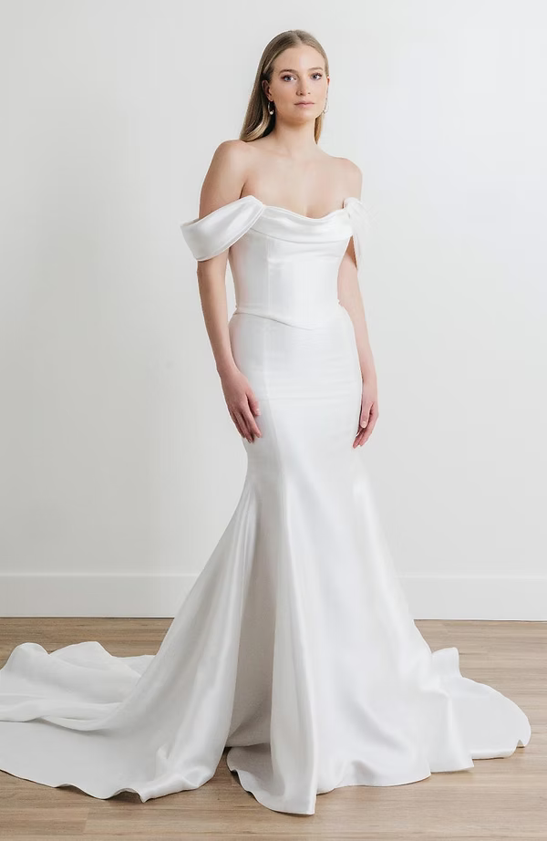 Woman in a white, off-the-shoulder mermaid wedding dress, standing in a studio.