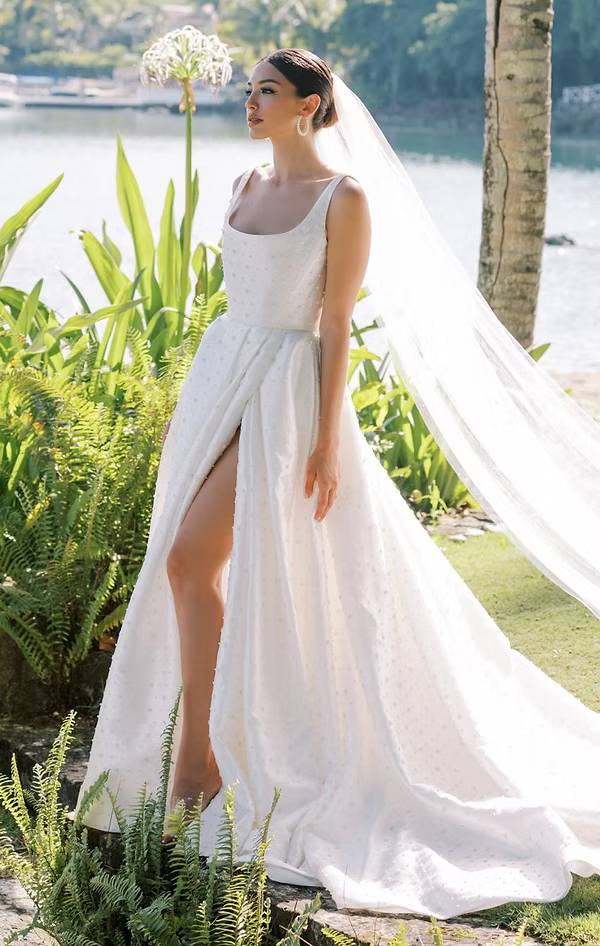 Bride in white gown with high slit, veil blowing, stands outside, water and greenery in background.