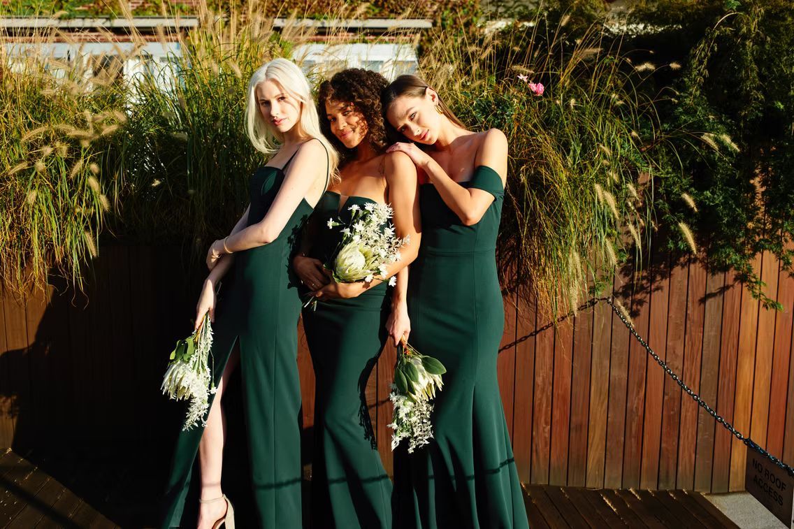 Three women in emerald green dresses hold bouquets, posing outdoors.