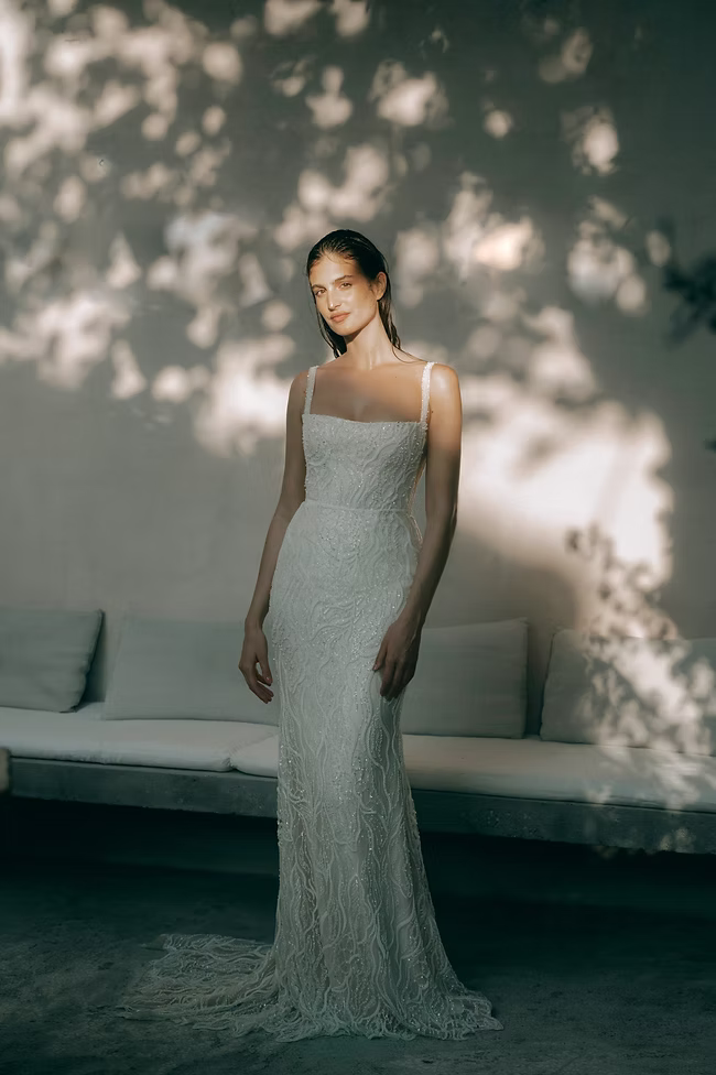 Woman in a white beaded wedding dress stands outside, sun dappled shadows on a white wall.