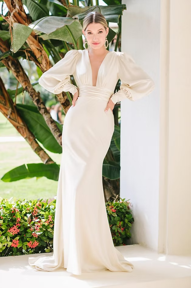 Woman in a white gown with plunging neckline and long sleeves posing outside.