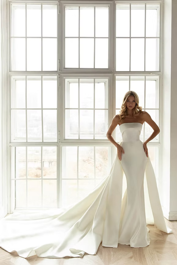 Woman in white strapless wedding dress with long train, standing by a large window.