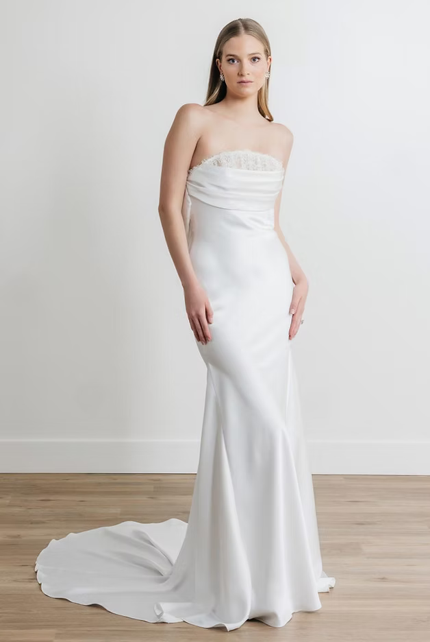Woman in a white strapless wedding gown with a flowing train, standing in front of a white wall.