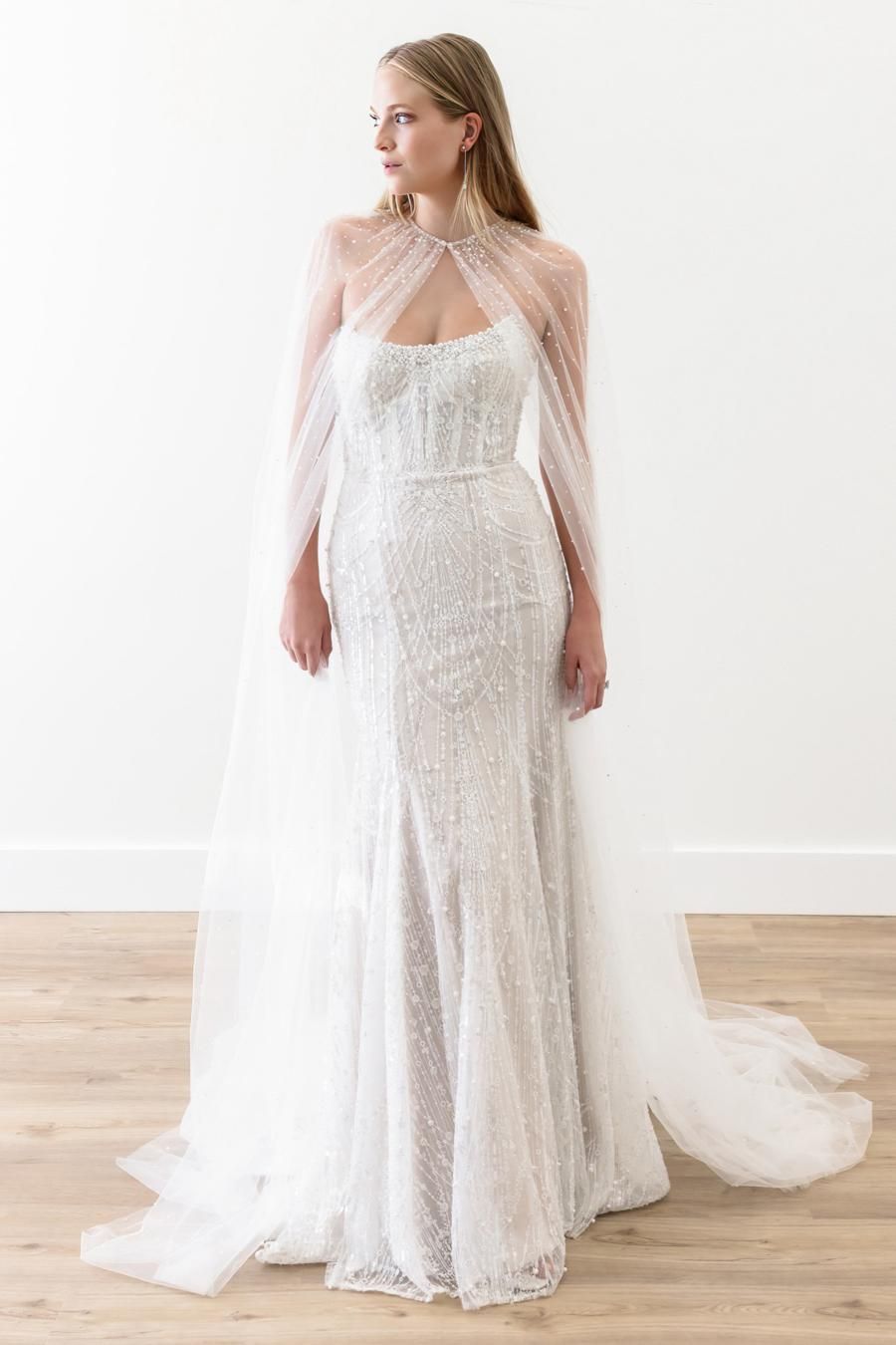 Woman in beaded wedding dress with cape, standing in front of a white wall.