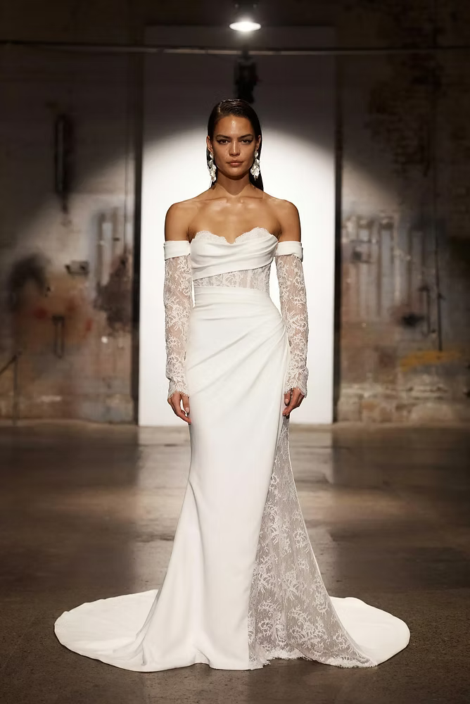 Woman in white off-the-shoulder gown with lace sleeves and a train; runway setting.