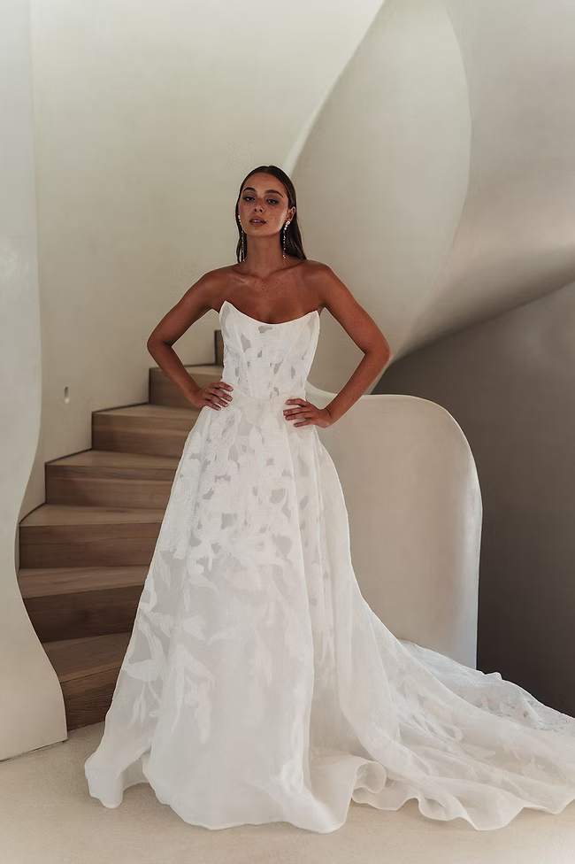 Woman in strapless white wedding dress poses on a spiral staircase, hands on hips, facing the camera.