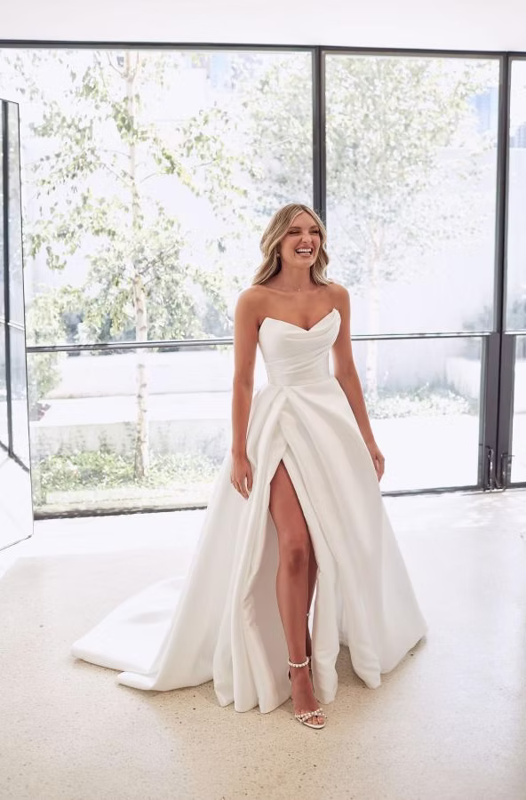 Woman in strapless white wedding dress with high slit, smiling, in a bright room.