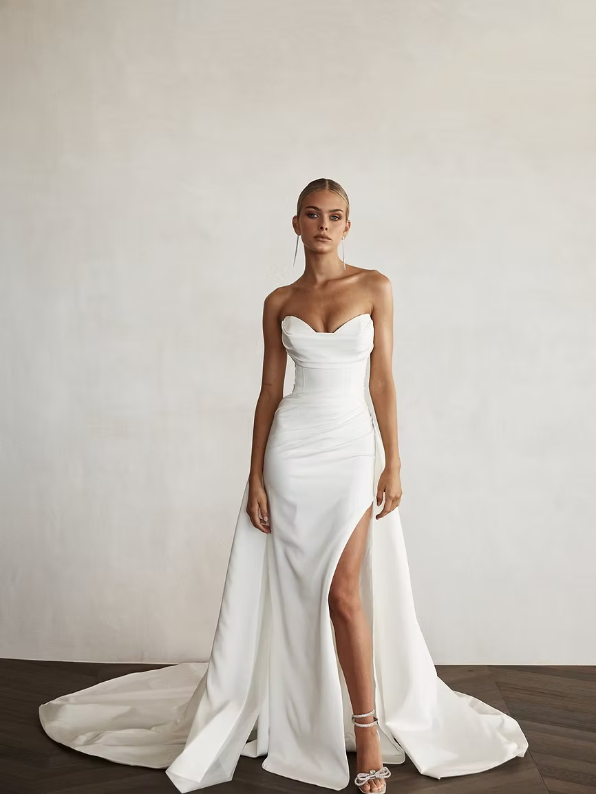 Woman in white strapless wedding dress with thigh-high slit and long train, standing in a room.