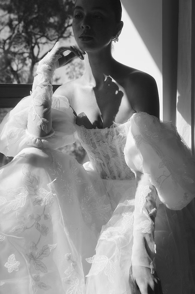 Woman in elaborate lace gown, bathed in sunlight, seated near a window, wearing long gloves.