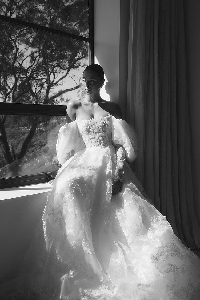 Woman in a white, layered gown sits by a window, sunlight illuminating the dress and a tree outside.