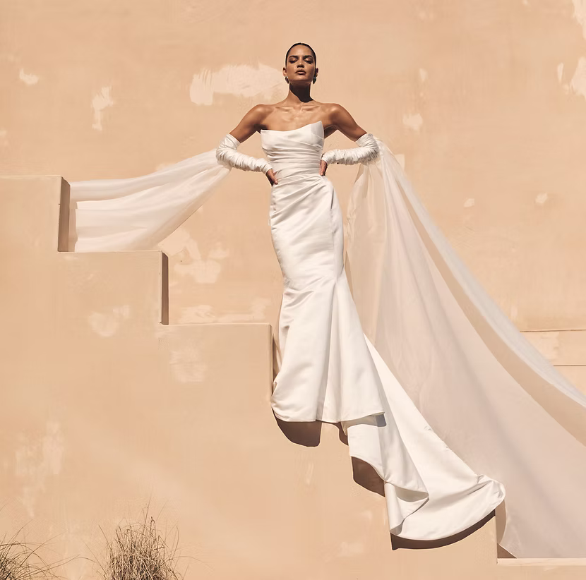 Woman in white strapless wedding gown with long train, posing on peach steps, wearing gloves.