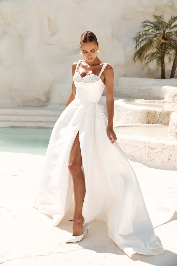 Woman in white wedding dress with high slit, standing by pool and white stone structures.