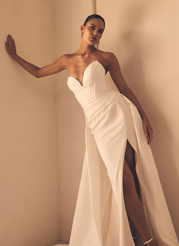 Woman in white strapless wedding gown with high slit, posing by a wall.