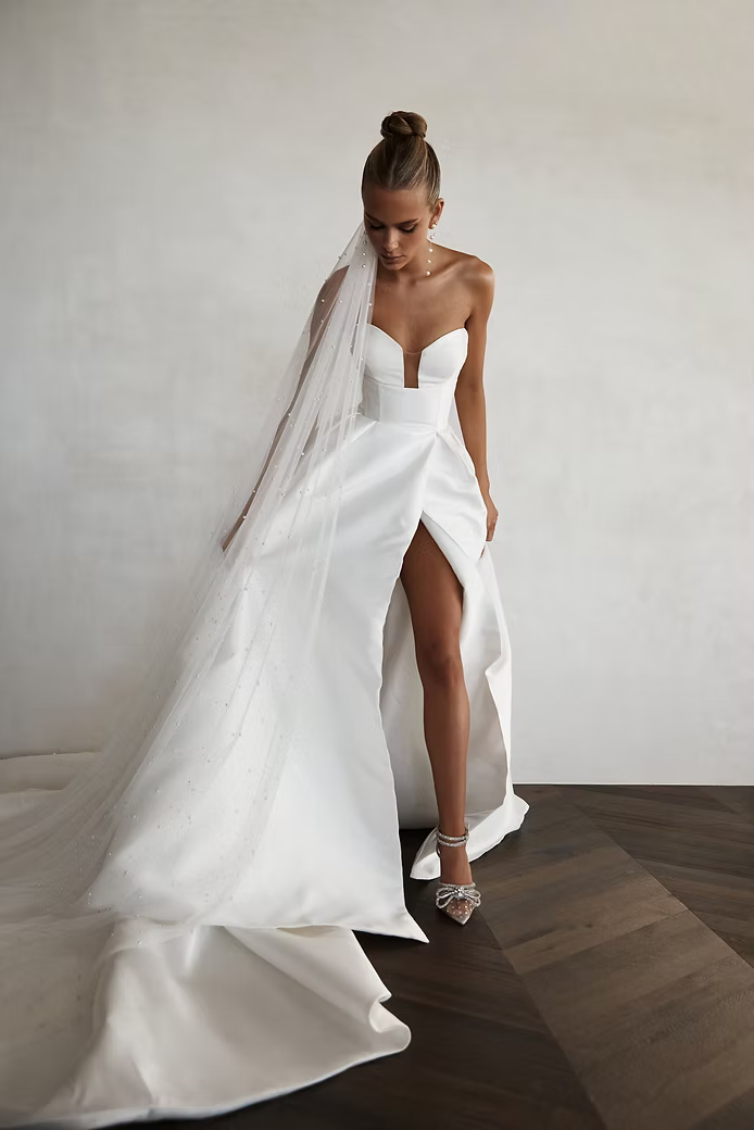 Bride in white strapless gown with high slit and veil, standing indoors on wood floor.