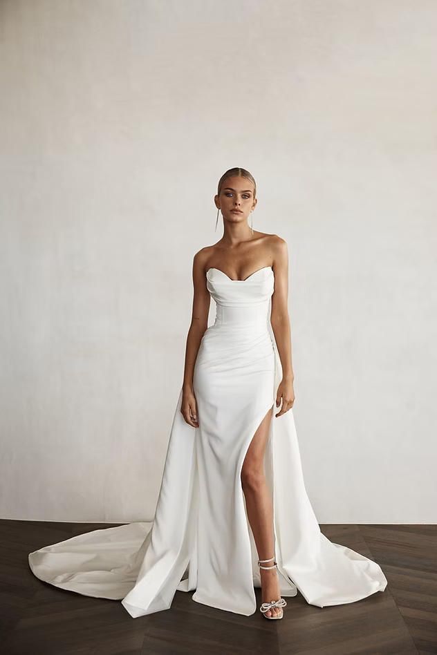 Woman in white strapless wedding dress with high slit and train, standing against a white wall.