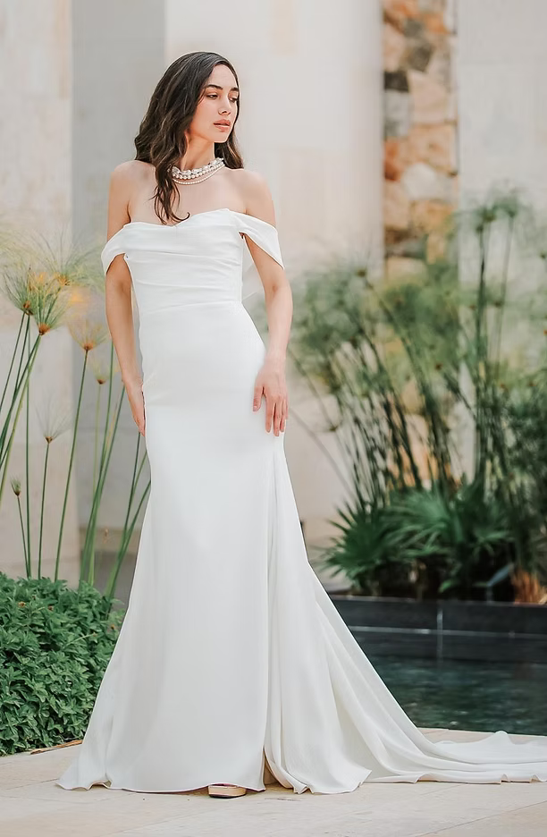 Woman in a white off-the-shoulder wedding dress, posing outdoors near water and greenery.