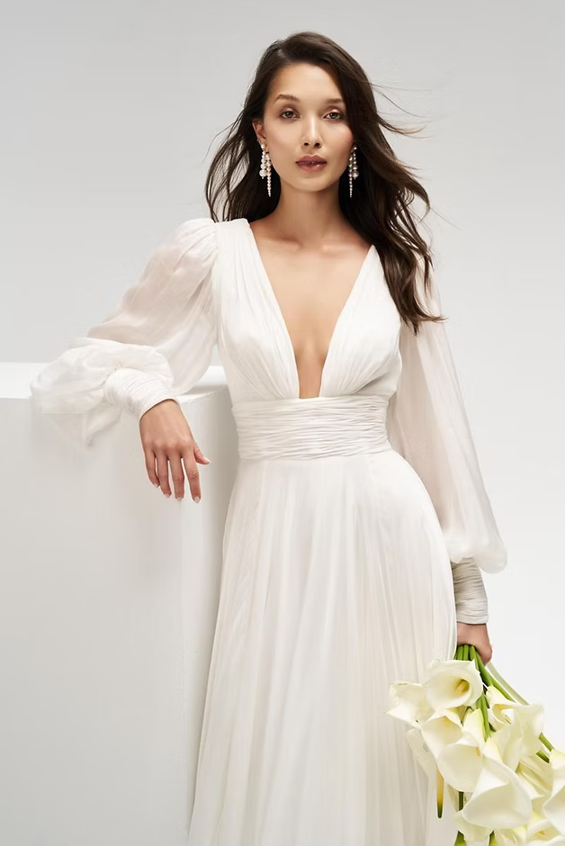 Woman in white wedding dress holding flowers, leaning against a white surface, looking forward.