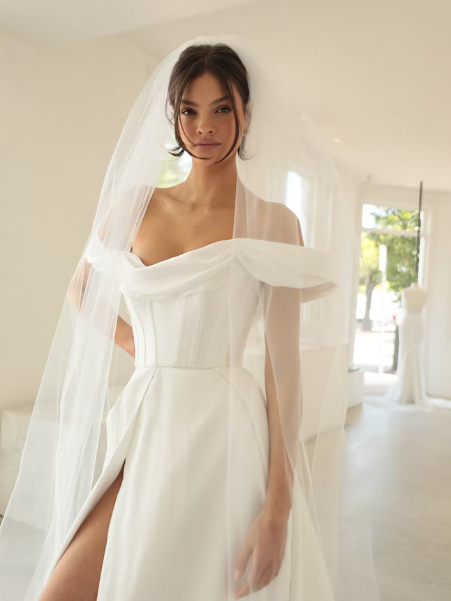 Woman in a white off-shoulder wedding dress and veil, standing indoors.