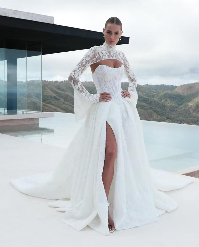 Woman in white wedding gown with a high slit, standing outdoors by a pool, arms on hips.