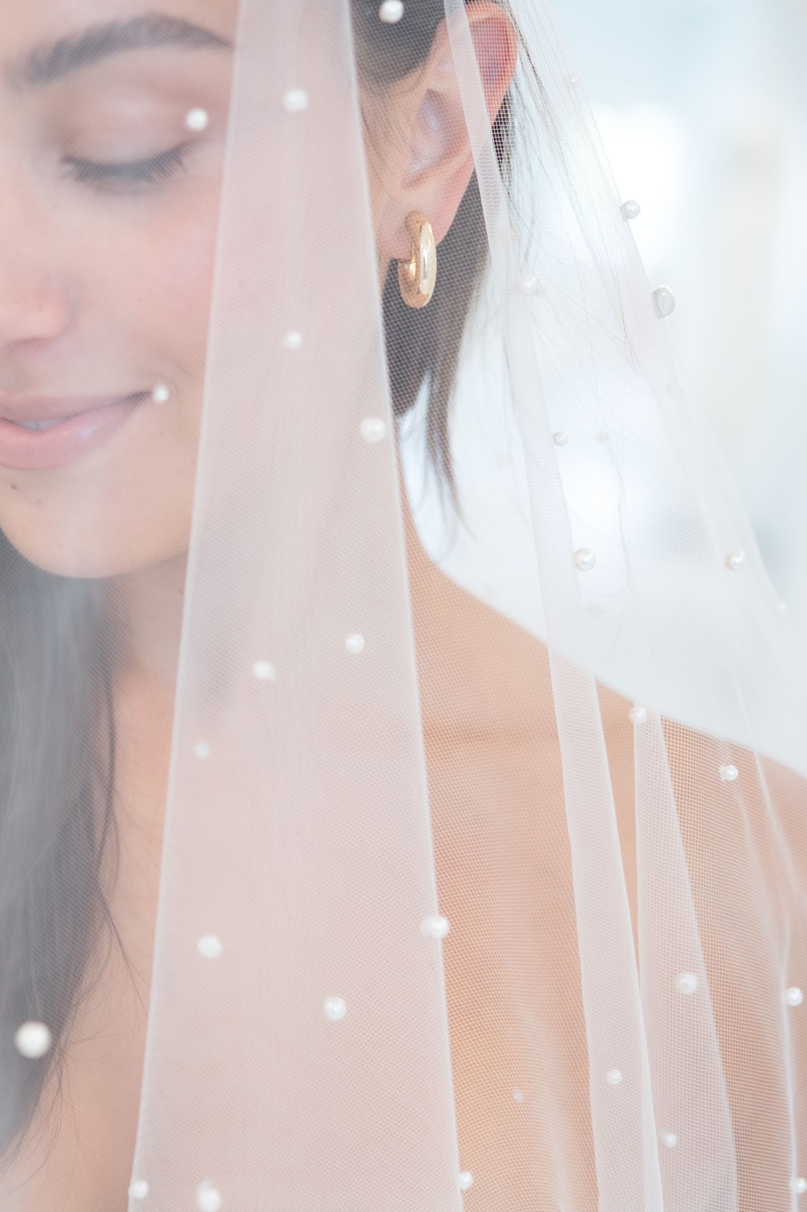 Woman with a pearl-studded veil, smiling with closed eyes, wearing gold hoop earrings.