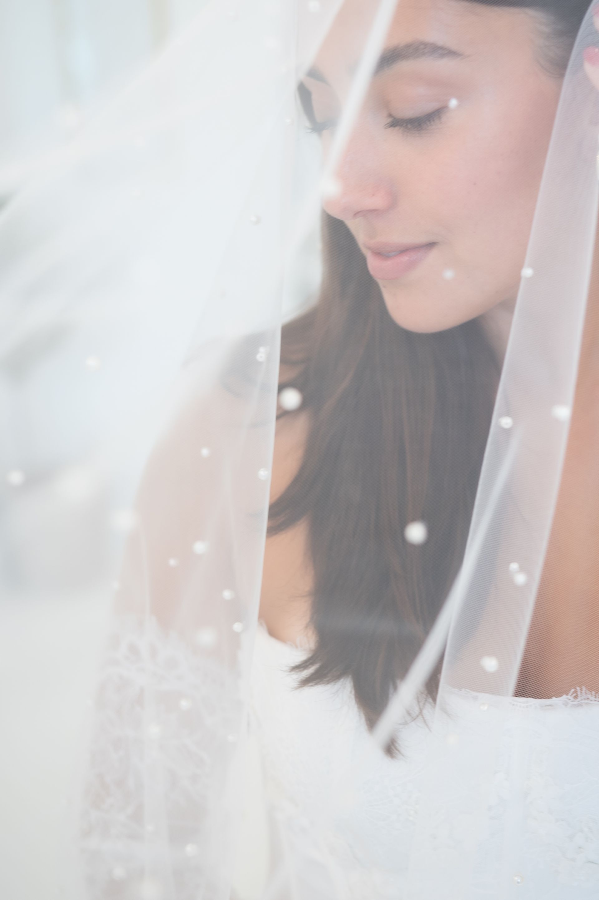 Bride with eyes closed, veiled in sheer white fabric adorned with small pearls.