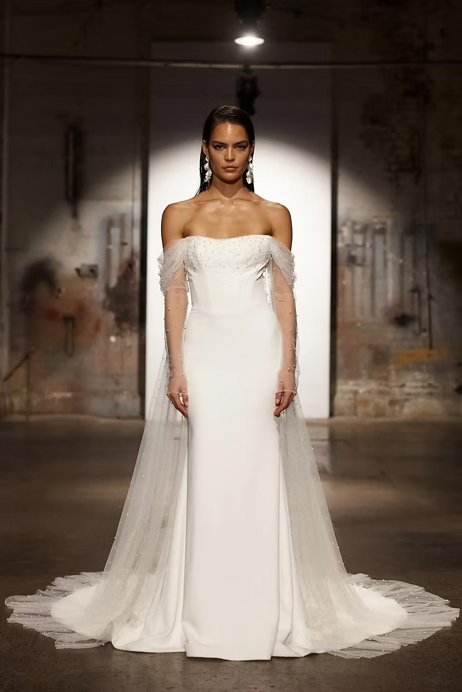Bride in white off-the-shoulder gown with sheer sleeves and train, walking runway.