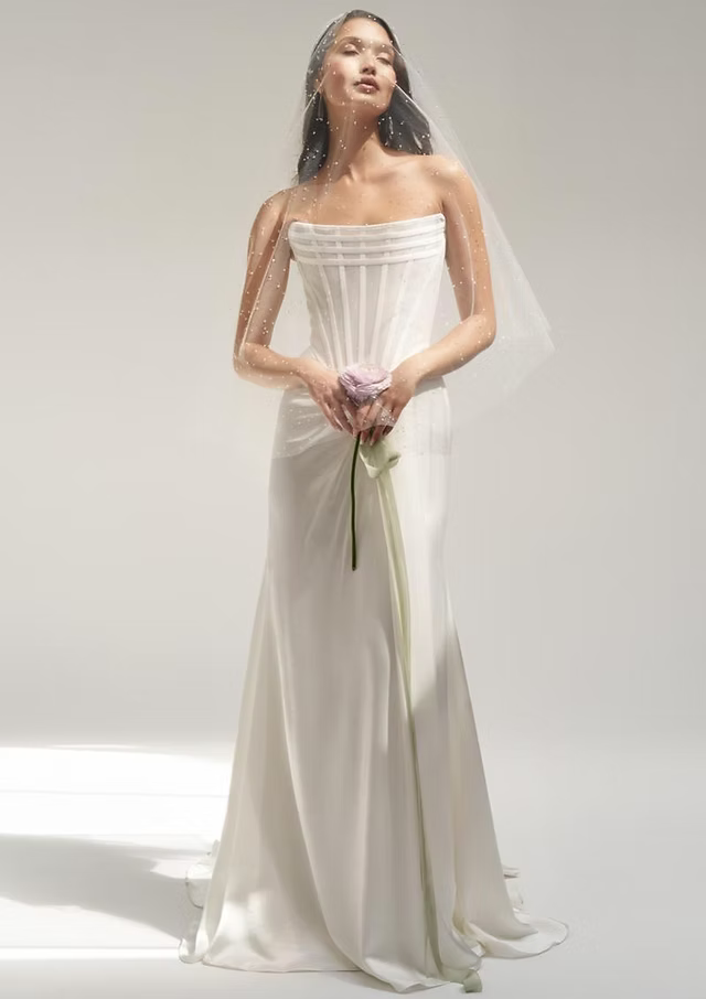 Woman in a strapless white wedding dress, holding a flower, with a veil, standing in a studio.